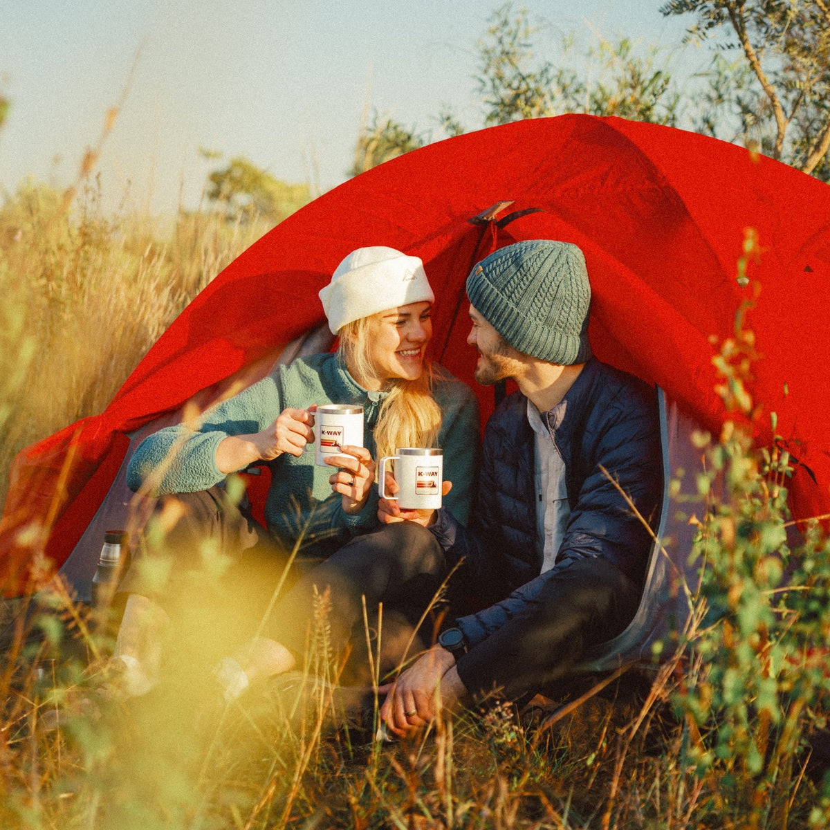 Two hikers smiling with coffee mugs in front of a red tent in golden grass